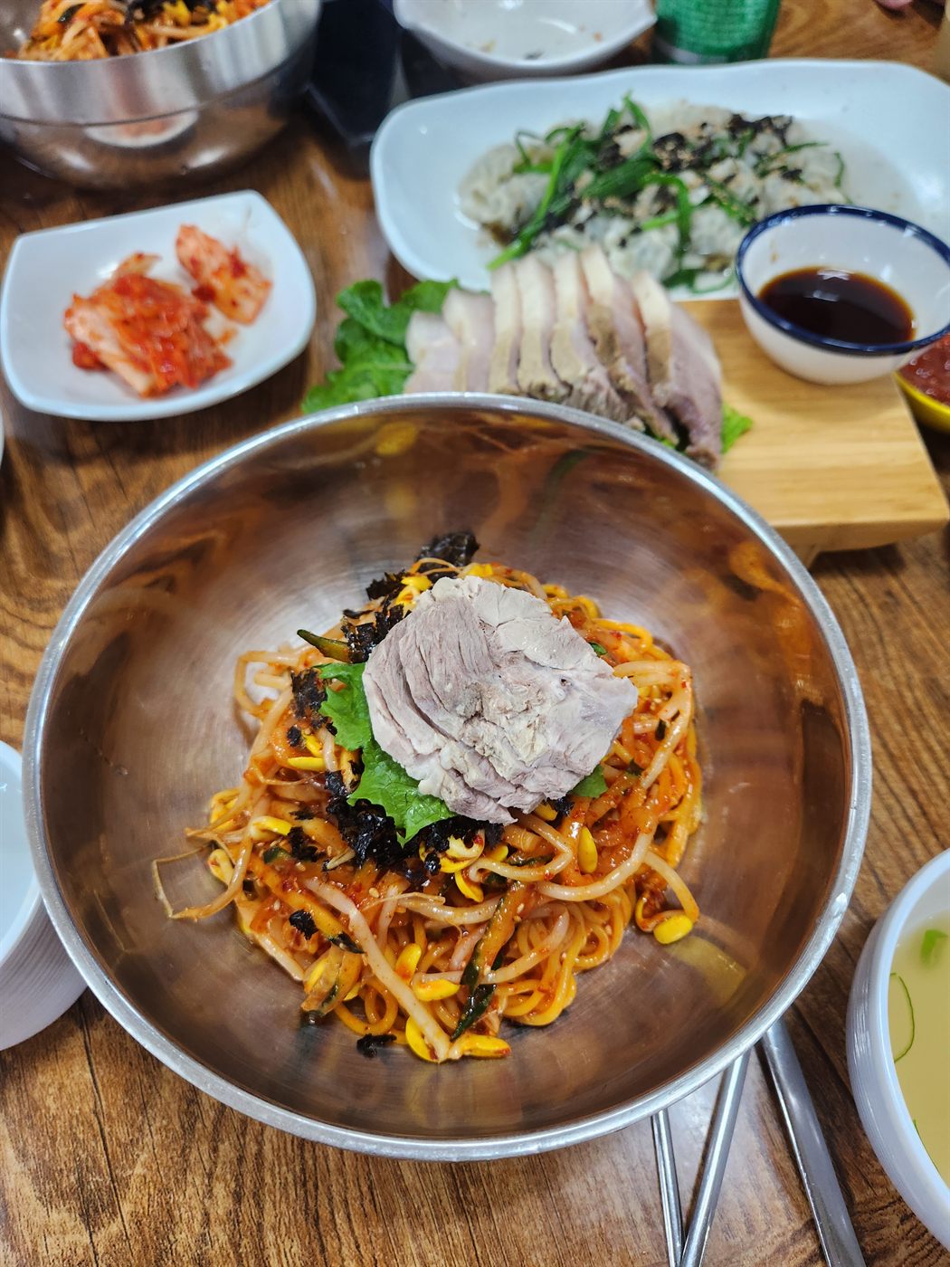 나무 식탁 위 스테인리스 대접에 담긴 비빔국수. 수육 한 점과 콩나물, 김이 올라가 있고 옆으로 수육 접시, 만두, 김치, 국물이 놓여 있다