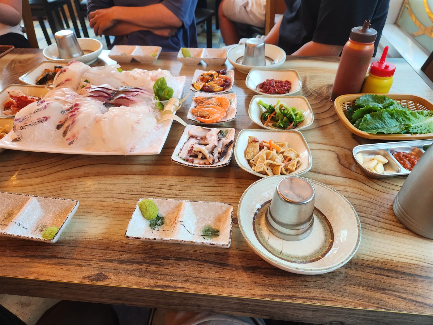 나무 식탁에 차려진 회 한 상. 흰 접시에 담긴 회를 가운데 두고 새우, 문어, 나물 등 밑반찬 여러 접시와 상추 바구니가 놓여 있다